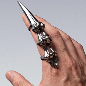 Puede incluir: Un anillo de dedo plateado con un diseño largo y puntiagudo. El anillo tiene una estructura detallada y texturizada que envuelve el dedo, con una punta afilada y alargada que se extiende desde la punta del dedo. El anillo es una joya única.