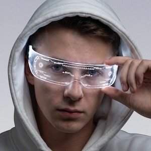 May include: Clear LED glasses with a futuristic design, emitting a white glow. The glasses have circuit-like patterns and are worn by a person in a white hooded top. The glasses have a digital display.