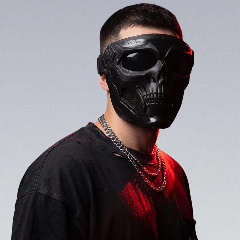 May include: A black skull mask with large eye openings and a detailed skeletal design. The mask is worn with a black shirt and a silver chain necklace. The background is a gradient of gray and red.