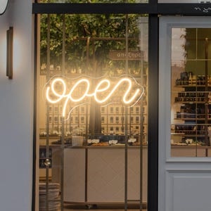 Pode incluir: Fachada de uma loja com um letreiro de néon "open" na vitrine. O letreiro está em uma fonte cursiva e emite uma luz quente. O interior da loja é visível através do vidro, com prateleiras e expositores.