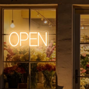 Pode incluir: Uma placa de néon numa montra exibe a palavra "OPEN" num brilho quente e convidativo. A janela está cheia de coloridos bouquets de flores, criando uma atmosfera acolhedora. O nome da loja, "The Neon Vintage", é visível na porta.