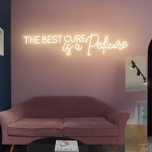 Könnte beinhalten: Ein rosa Samtsofa und ein schwarzer runder Couchtisch stehen vor einer rosa Wand. Ein Neon-Schild über dem Sofa lautet "THE BEST CURE is a Pedicure". Ein großer Spiegel und weiße Regale sind ebenfalls zu sehen.