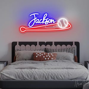 Op de afbeelding: Een neonbord in een slaapkamer toont de naam "Jackson" in witte letters, boven een honkbal- en knuppelontwerp. Het bord gloeit met blauw en rood licht. De slaapkamer heeft een bed met grijs beddengoed en decoratieve kussens.