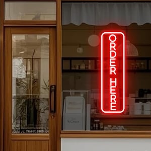 May include: A rectangular neon sign with a bright red border and the text "ORDER HERE" in white capital letters, mounted on the interior of a glass storefront window next to a wooden door with a glass panel and a dark metal handle.