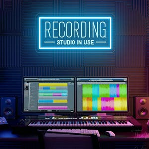 May include: A rectangular neon sign with a bright blue light displaying the text RECORDING STUDIO IN USE in a clean sans-serif font. The sign is mounted on a dark wall covered in acoustic foam panels above a professional music studio setup.