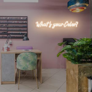 May include: A neon sign with the text What's your Color? in a cursive font mounted on a pink wall above a wooden nail salon desk with a grey side panel, a green leaf-patterned chair, a black lamp, and a wall-mounted nail polish display rack.