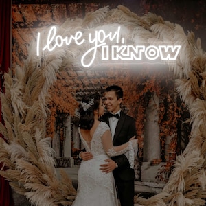 Könnte beinhalten: Ein Paar umarmt sich vor einem dekorativen Torbogen. Der Bogen ist mit getrocknetem Pampasgras und einem Neonschild mit der Aufschrift "I love you, I know" geschmückt. Die Braut trägt ein weißes Spitzenhochzeitskleid, der Bräutigam einen schwarzen Smoking.