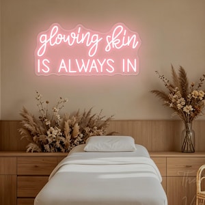 May include: A pink neon sign mounted on a wall featuring the cursive text glowing skin and the block letter text IS ALWAYS IN. The sign emits a soft light, creating a glow against the wall surface behind the illuminated lettering.