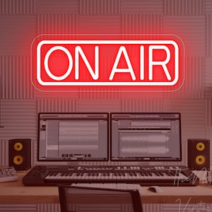 以下が含まれることがあります： 白い長方形のフレームに入った赤いネオンの「ON AIR」サインが机の上に吊り下げられています。2台のコンピューターモニターには、オーディオ編集ソフトウェアが表示されています。キーボード、スピーカー、その他のオーディオ機器が机の上にあり、レコーディングスタジオを連想させます。