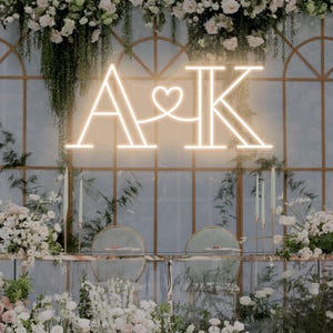 May include: A glowing neon sign featuring the letters A and K connected by a heart symbol. The sign emits a warm white light and is mounted on a decorative arched frame adorned with white flowers and greenery above a table with two chairs.