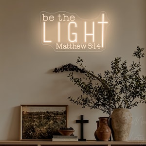 May include: A warm white neon sign mounted on a wall featuring the text "be the LIGHT" with a cross integrated into the letter T, followed by the text "Matthew 5:14" below. The sign emits a soft glow, illuminating the wall behind it.