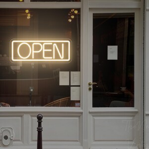 Könnte beinhalten: Ein rechteckiges Neonschild mit warmweißem Licht, das das Wort OPEN in einer klaren serifenlosen Schrift anzeigt. Das Schild ist im Glasfenster eines weißen Ladenlokals montiert und verfügt über einen dünnen Rahmen, der den Bereich beleuchtet.