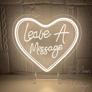 Op de afbeelding: Een wit neon hartvormig bord met de tekst "Leave A Message" in een cursief lettertype. Het bord is gemonteerd op een transparante standaard. De achtergrond is een getextureerde muur.