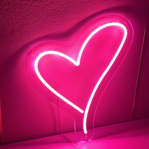 May include: A bright pink neon heart-shaped light fixture. The light is outlined in a bright white, creating a glowing effect against the pink background. The fixture is mounted on a pink wall.