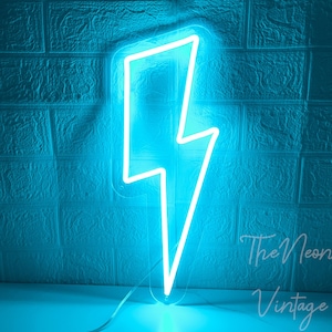 Könnte beinhalten: Ein hellblaues Neon-Blitzzeichen vor einer strukturierten blauen Backsteinmauer. Das Schild hat eine stilisierte Zickzackform mit dem Text "The Neon Vintage" in der unteren rechten Ecke. Das Schild leuchtet und scheint aus Glas zu sein.