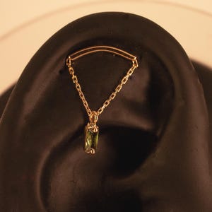 May include: A gold ear piercing jewellery piece featuring a curved bar connected by two delicate chains to a dangling rectangular green gemstone. The jewellery is displayed on a dark ear-shaped model, showcasing the chain drape and the gemstone.