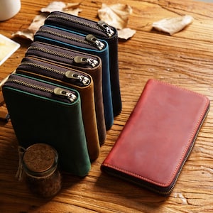 May include: A collection of leather wallets in various colors, including green, brown, blue, and red. Each wallet features a zipper closure with a brass-colored pull. The wallets are arranged on a wooden surface.