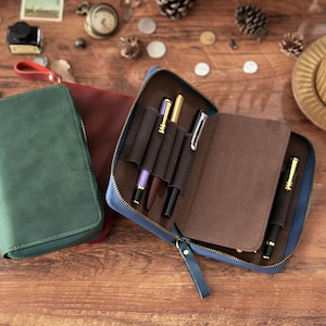 Peut inclure: Un étui à stylos en cuir bleu ouvert, révélant plusieurs stylos dans des supports marron. Un étui en cuir vert fermé et un étui en cuir bordeaux fermé sont également visibles. Les stylos sont dorés, noirs et violets.