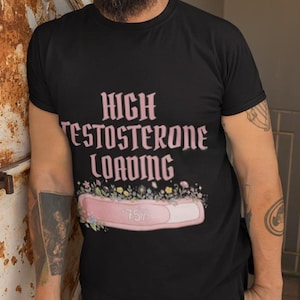 Puede incluir: Camiseta negra con el texto rosa "HIGH TESTOSTERONE LOADING". Debajo, una barra de carga rosa con "75%" y detalles florales. La camiseta la lleva una persona.