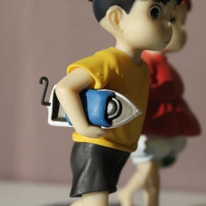 May include: A figurine of a character wearing a yellow t-shirt and black shorts, holding a blue and white iron under one arm. A second figurine wearing a red top and white shorts is partially visible in the background behind the first figure.