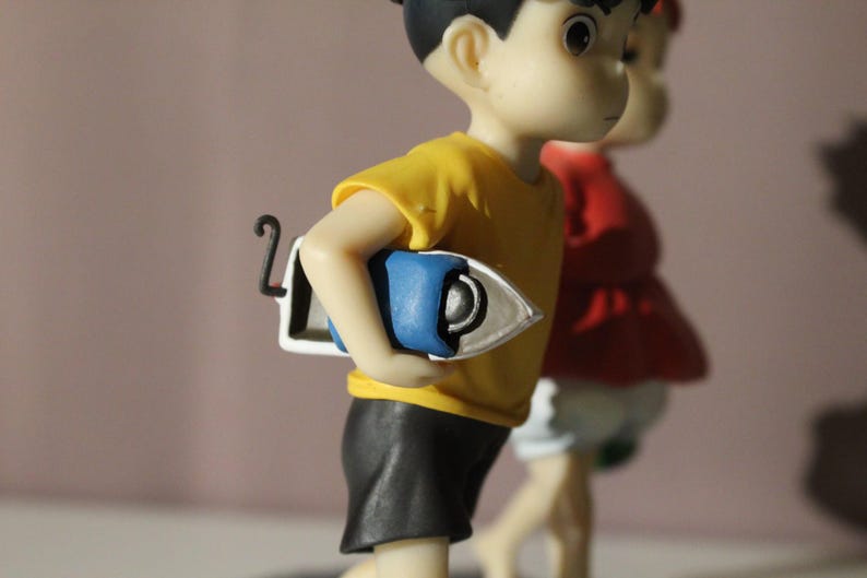 May include: A figurine of a character wearing a yellow t-shirt and black shorts, holding a blue and white iron under one arm. A second figurine wearing a red top and white shorts is partially visible in the background behind the first figure.