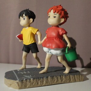 May include: A figurine featuring two characters walking hand-in-hand on a gray textured base. One character has black hair, a yellow shirt, and black shorts, holding a red object. The other has red hair, a red top, and white shorts, carrying a green bucket.
