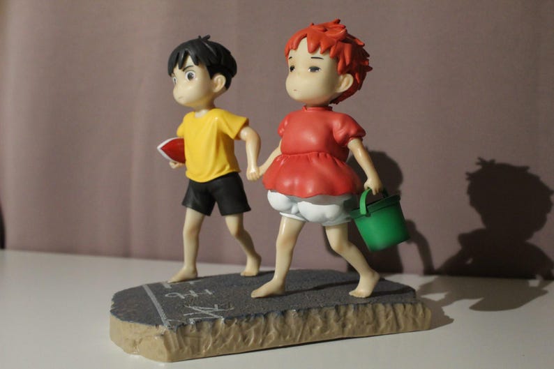 May include: A figurine featuring two characters walking hand-in-hand on a gray textured base. One character has black hair, a yellow shirt, and black shorts, holding a red object. The other has red hair, a red top, and white shorts, carrying a green bucket.