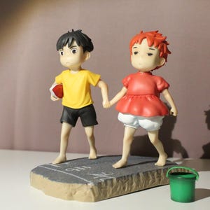 May include: A figurine featuring two characters holding hands, one with black hair wearing a yellow shirt and black shorts, the other with red hair wearing a red top and white shorts, standing on a gray base with a small green bucket nearby.