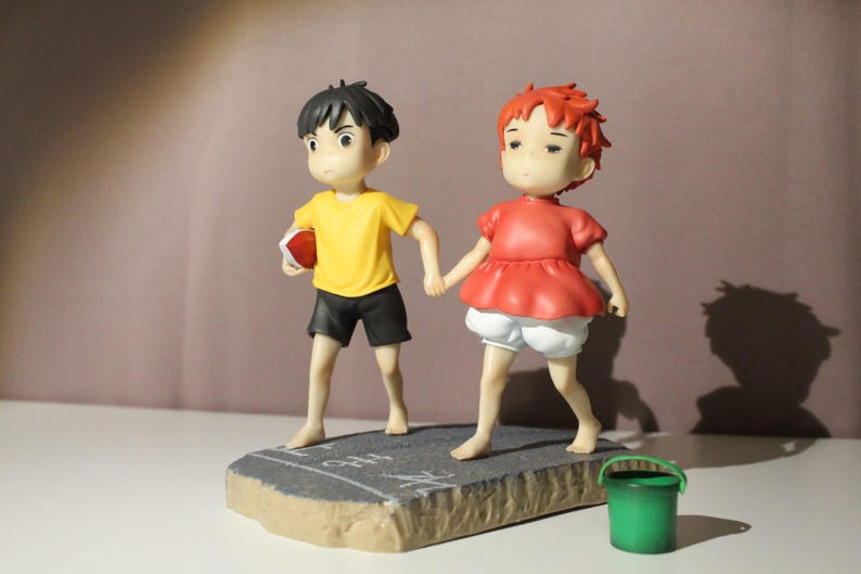 May include: A figurine featuring two characters holding hands, one with black hair wearing a yellow shirt and black shorts, the other with red hair wearing a red top and white shorts, standing on a gray base with a small green bucket nearby.