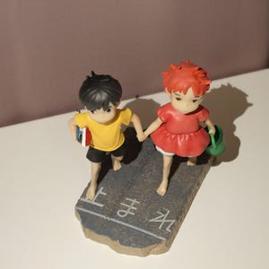 May include: A figurine featuring two children holding hands, standing on a gray base with white Japanese text. The child on the left wears a yellow shirt and black shorts, while the child on the right wears a red dress and carries a green bucket.
