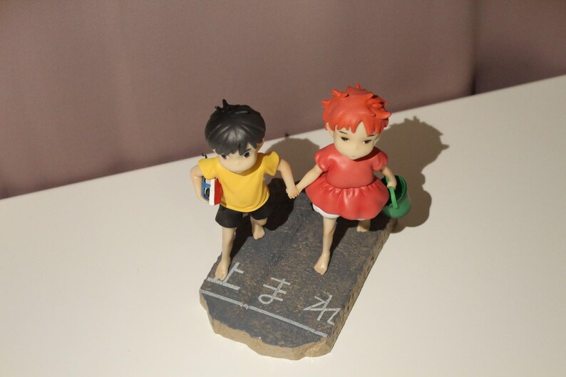May include: A figurine featuring two children holding hands, standing on a gray base with white Japanese text. The child on the left wears a yellow shirt and black shorts, while the child on the right wears a red dress and carries a green bucket.