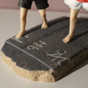 May include: A figurine base featuring a textured dark gray surface with white painted lines and the text "H" and "M" visible. The base supports two figures, one wearing black shorts and the other wearing white shorts, both with bare feet.