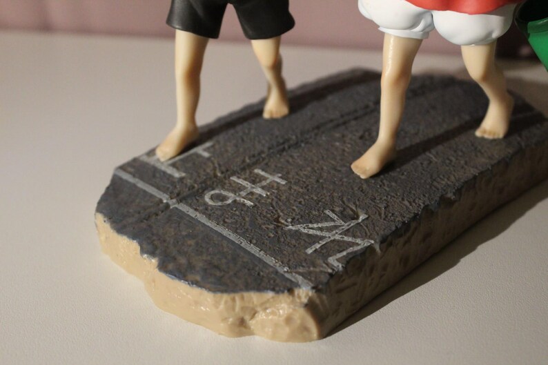 May include: A figurine base featuring a textured dark gray surface with white painted lines and the text "H" and "M" visible. The base supports two figures, one wearing black shorts and the other wearing white shorts, both with bare feet.