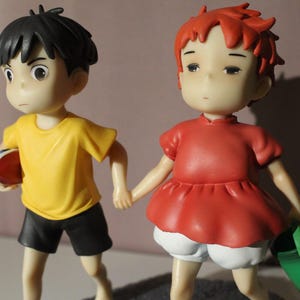 May include: Two small figurines holding hands. The figure on the left has black hair, a yellow shirt, and black shorts, holding a red object. The figure on the right has short red hair, a red dress, white shorts, and carries a small green bucket.