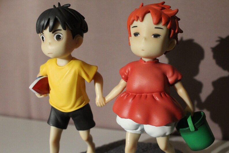 May include: Two small figurines holding hands. The figure on the left has black hair, a yellow shirt, and black shorts, holding a red object. The figure on the right has short red hair, a red dress, white shorts, and carries a small green bucket.
