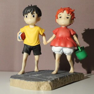 May include: A figurine featuring two characters holding hands. One character wears a yellow shirt and black shorts, holding a red and white object. The other character has red hair, a red top, and white shorts, carrying a small green bucket.
