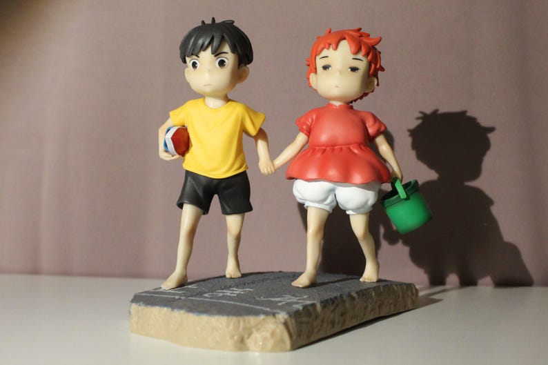 May include: A figurine featuring two characters holding hands. One character wears a yellow shirt and black shorts, holding a red and white object. The other character has red hair, a red top, and white shorts, carrying a small green bucket.