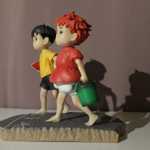 May include: A figurine featuring two characters walking on a textured gray base. One character has short red hair, a red shirt, and white shorts, carrying a green bucket. The other character has short black hair, a yellow shirt, and black shorts.