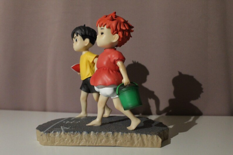 May include: A figurine featuring two characters walking on a textured gray base. One character has short red hair, a red shirt, and white shorts, carrying a green bucket. The other character has short black hair, a yellow shirt, and black shorts.