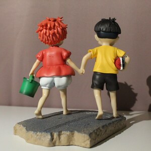 May include: A figurine of two figures holding hands, standing on a textured gray base. The figure on the left has short red hair, a red shirt, white shorts, and carries a green bucket. The figure on the right has black hair, a yellow shirt, and black shorts.