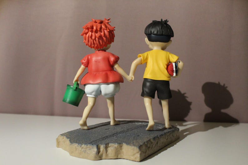 May include: A figurine of two figures holding hands, standing on a textured gray base. The figure on the left has short red hair, a red shirt, white shorts, and carries a green bucket. The figure on the right has black hair, a yellow shirt, and black shorts.