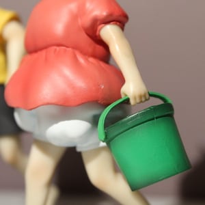 May include: A figurine of a character wearing a red shirt and white shorts, holding a small green plastic bucket. In the background, a second figurine is partially visible, wearing a yellow shirt and black shorts, with light skin tones.
