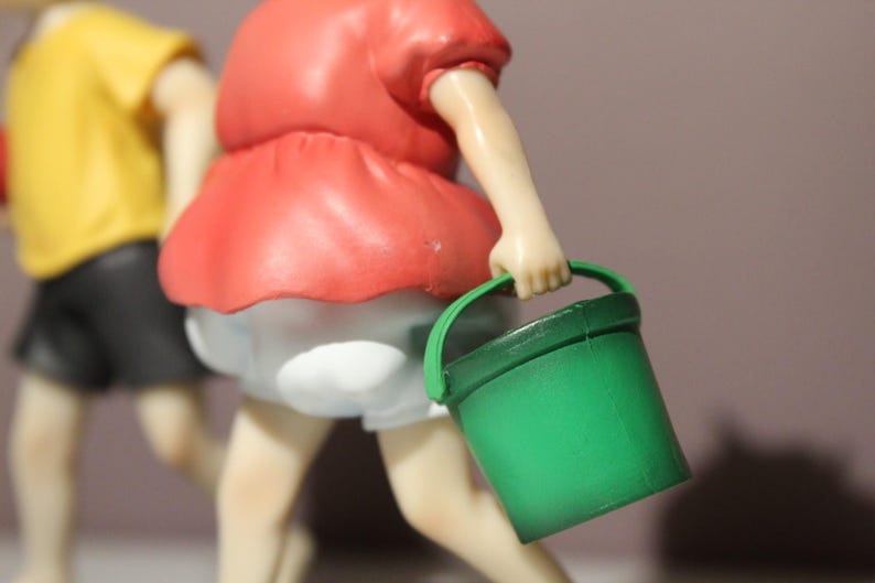 May include: A figurine of a character wearing a red shirt and white shorts, holding a small green plastic bucket. In the background, a second figurine is partially visible, wearing a yellow shirt and black shorts, with light skin tones.