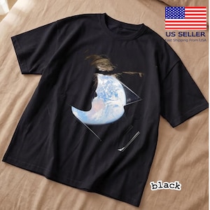 以下が含まれることがあります： 地球の描写と融合した人物のシルエットのグラフィックデザインが特徴の黒いTシャツ。デザインには、幾何学的な形の中に青と白の惑星が含まれています。「black」の文字が下部に印刷されています。