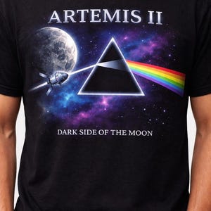 May include: Black t-shirt featuring a graphic design with the text "ARTEMIS II" above a black triangle, a rainbow, and a space scene. The words "DARK SIDE OF THE MOON" are below the graphic.