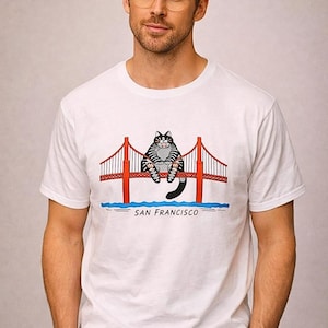 May include: White t-shirt featuring a cartoon cat sitting on the Golden Gate Bridge in San Francisco. The bridge is red with white accents, and the text "SAN FRANCISCO" is below the bridge.