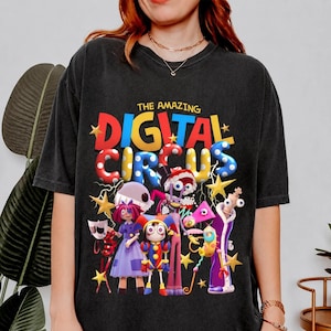 May include: Black t-shirt featuring the text "THE AMAZING DIGITAL CIRCUS" in colorful bubble letters, with cartoon characters and stars. The design includes a variety of whimsical figures in vibrant colors, creating a playful and eye-catching graphic.