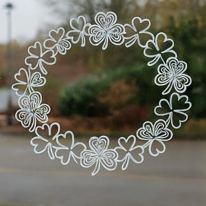 May include: A white shamrock wreath design on a glass surface. The wreath is composed of stylized four-leaf clovers, creating a circular pattern. The background shows a blurred outdoor scene with trees and a cloudy sky.