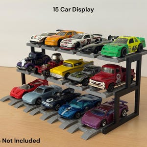 May include: A black three-tiered display stand with gray tracks holding various die-cast toy cars. The stand features the text 15 Car Display at the top and Cars Not Included at the bottom. The structure is designed to organize and showcase small vehicles.