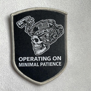 Könnte beinhalten: Ein schwarzer, schildförmiger Aufnäher mit hellgrauem Rand. Das Design zeigt einen weißen Totenkopf mit taktischem Helm, kleiner amerikanischer Flagge und Nachtsichtgerät. Der Text lautet OPERATING ON MINIMAL PATIENCE.